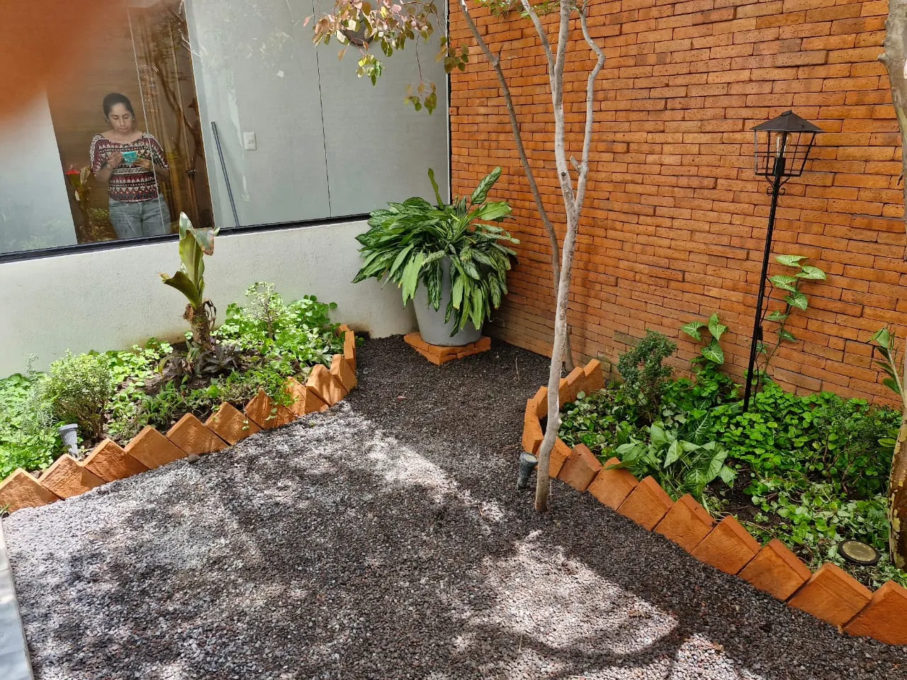Patio interior con jardineras de borde de barro, grava y farol junto a un muro de ladrillo